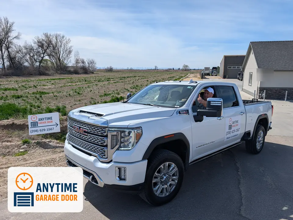 White Anytime Garage Door pickup with a driver giving a thumbs-up, next to a company yard sign by a field.