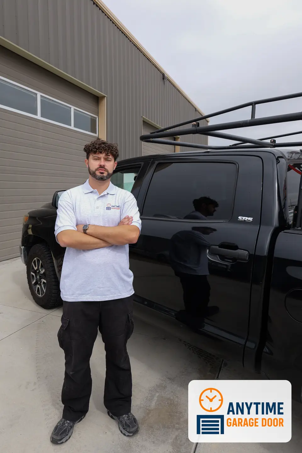 Technician in an Anytime Garage Door polo stands with arms crossed beside a black pickup with a ladder rack.