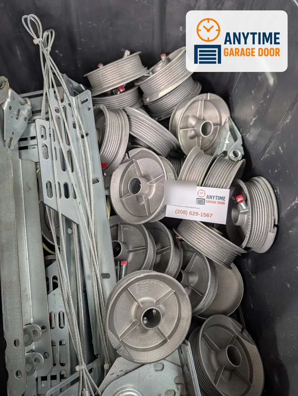 A bin of garage door cable drums, lift cables and steel brackets, with an Anytime Garage Door business card on top.