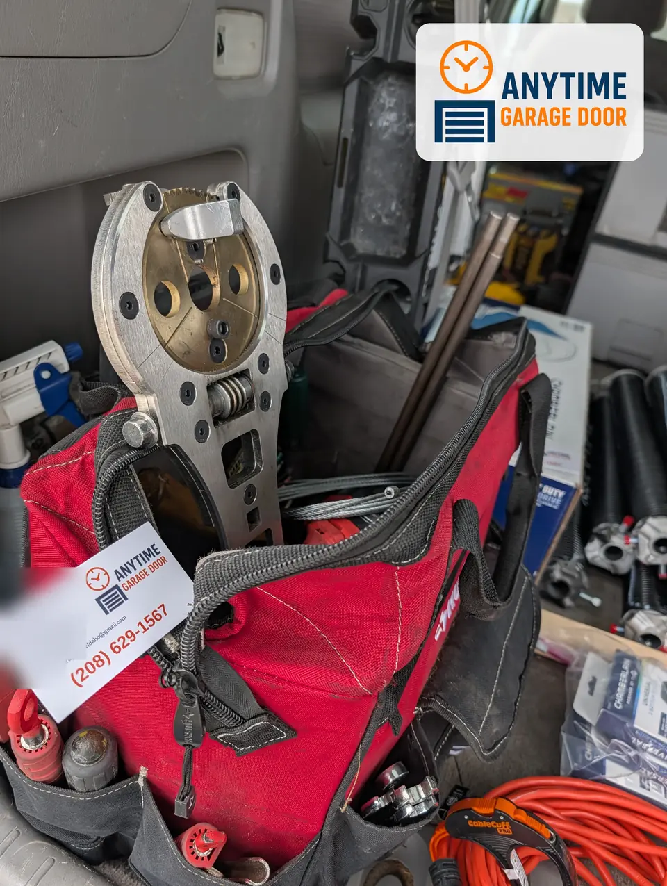 Metal hand tool, steel rods and coiled cable in a red tool bag, with an Anytime Garage Door business card in front.