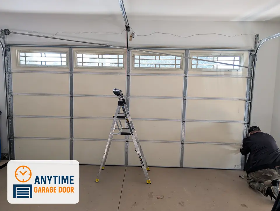 Inside view of a garage door with top windows, a step ladder in front and a worker kneeling at the right-side track.