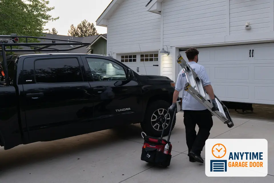 Technician carries a ladder on his shoulder and rolls a tool bag past a black pickup toward a white garage door.