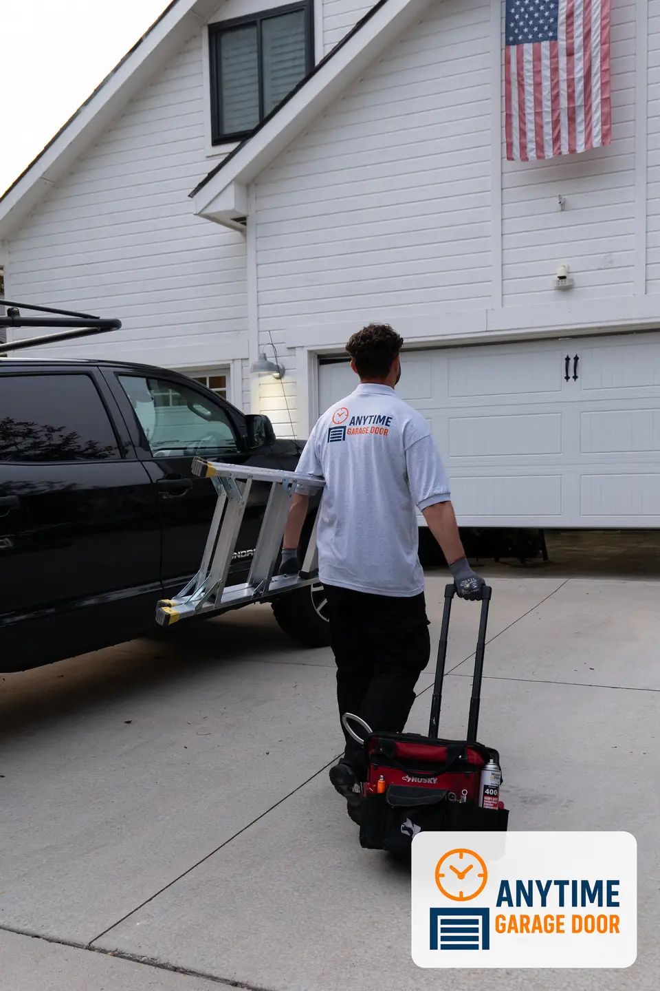 Technician carries a ladder under one arm and pulls a rolling tool bag across a driveway toward a garage door.