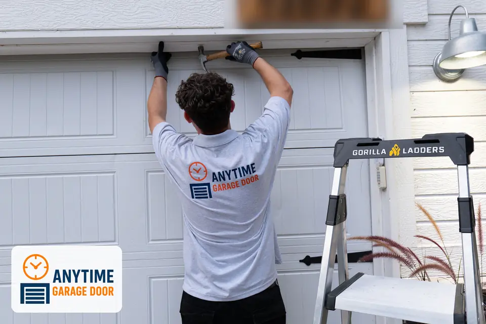 Technician uses a hammer along the top trim of a white garage door, with a step ladder beside him.