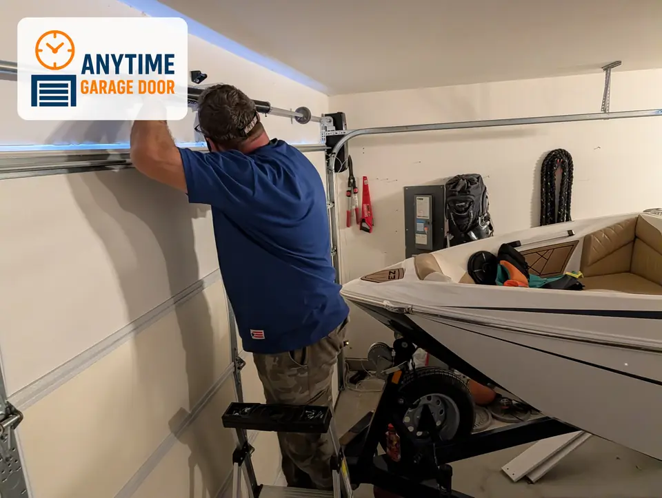 Worker on a stepladder gripping a torsion spring above a garage door, with a boat on a trailer in the garage