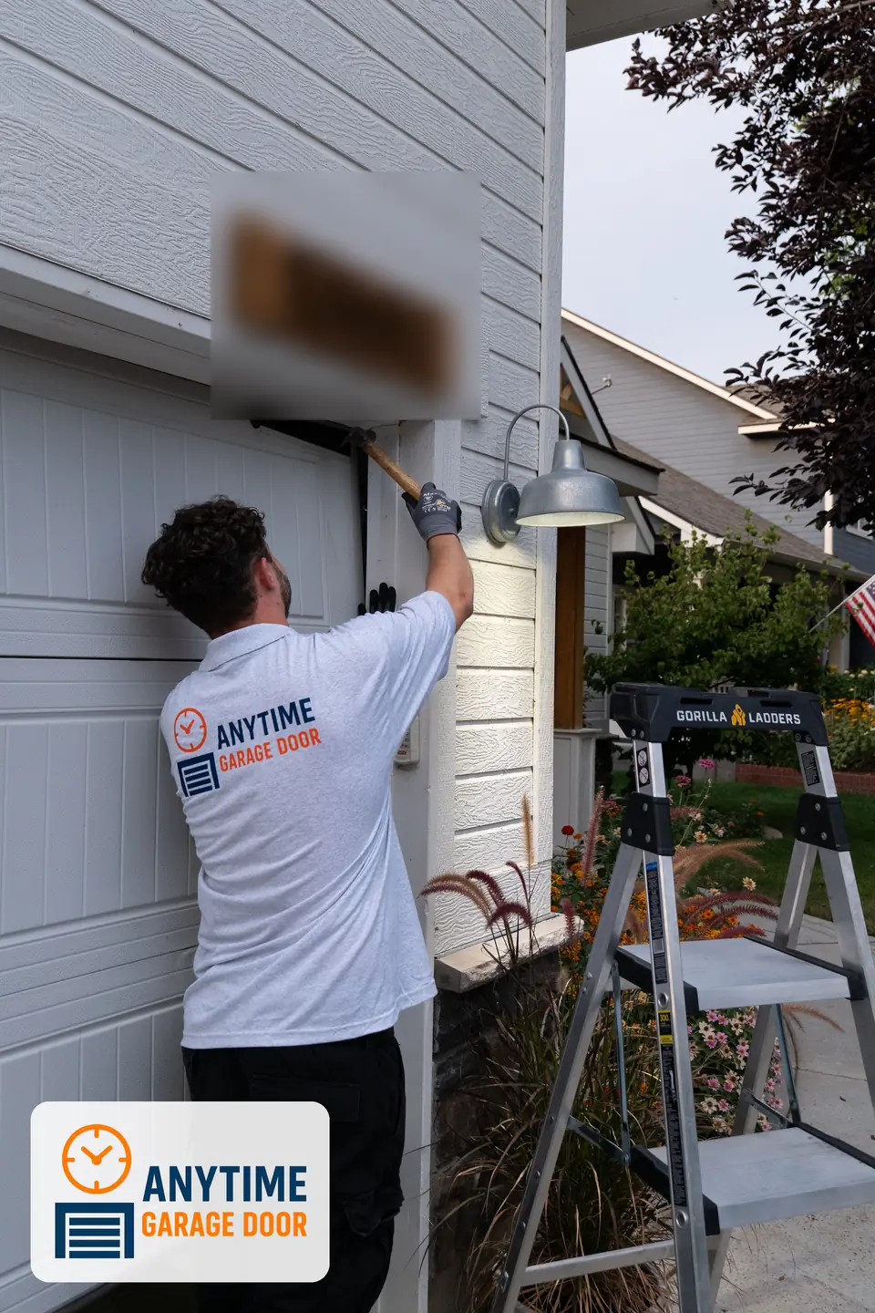 Technician hammers the trim at the top corner of a garage door opening, with a step ladder and lamp nearby.