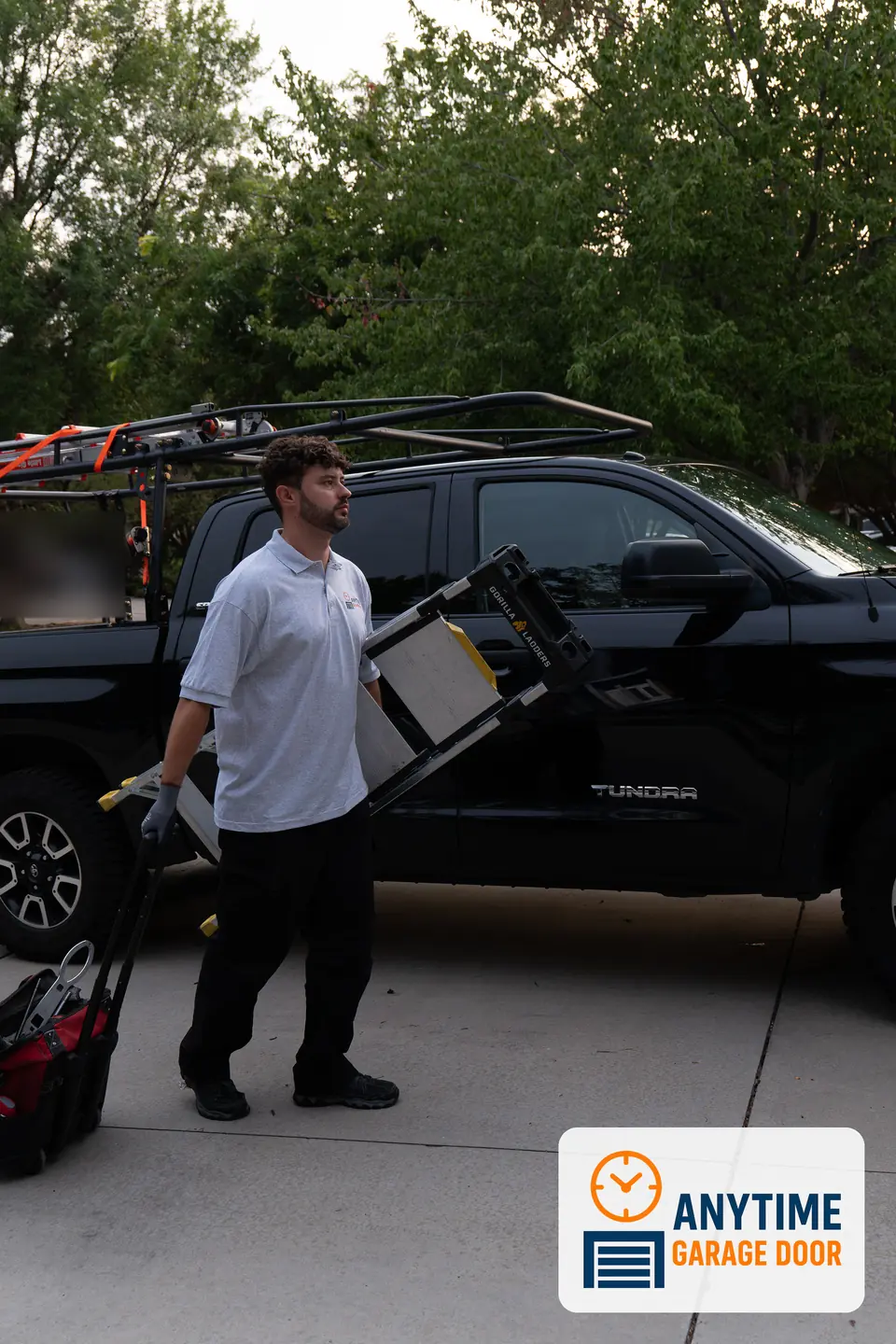 Technician lifts a step ladder from a black pickup with a ladder rack, with a rolling tool bag beside him.