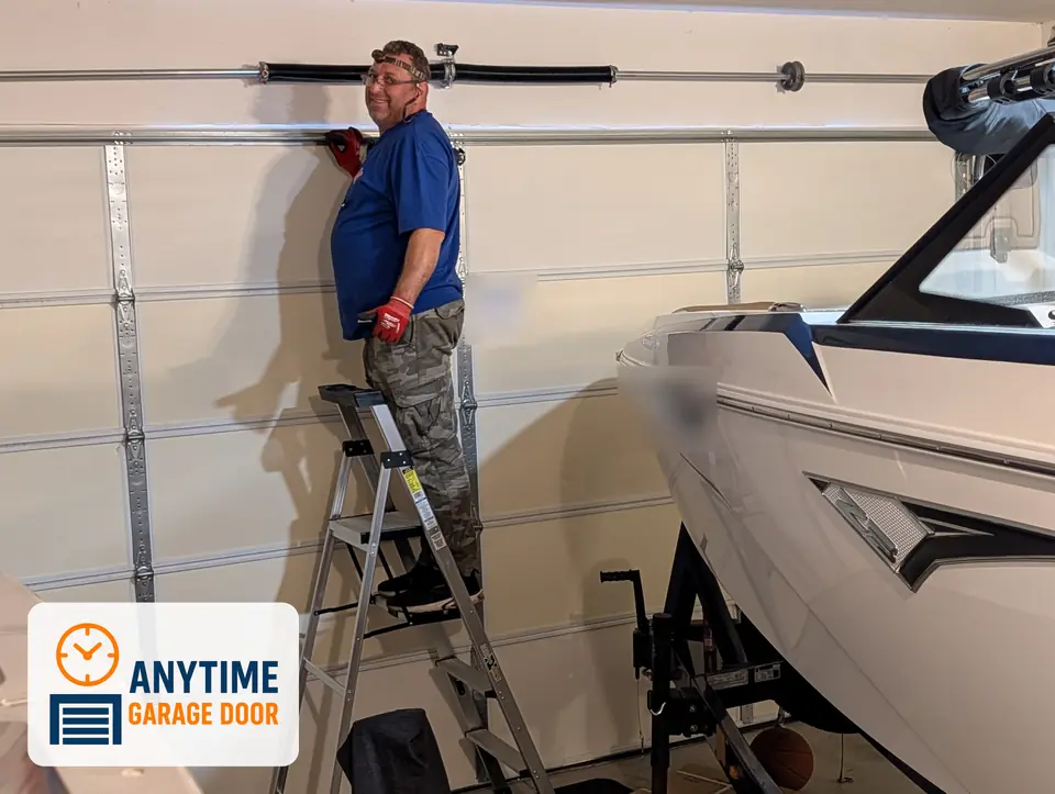Smiling man on a stepladder beside a torsion spring above a white garage door, with a boat parked in the garage