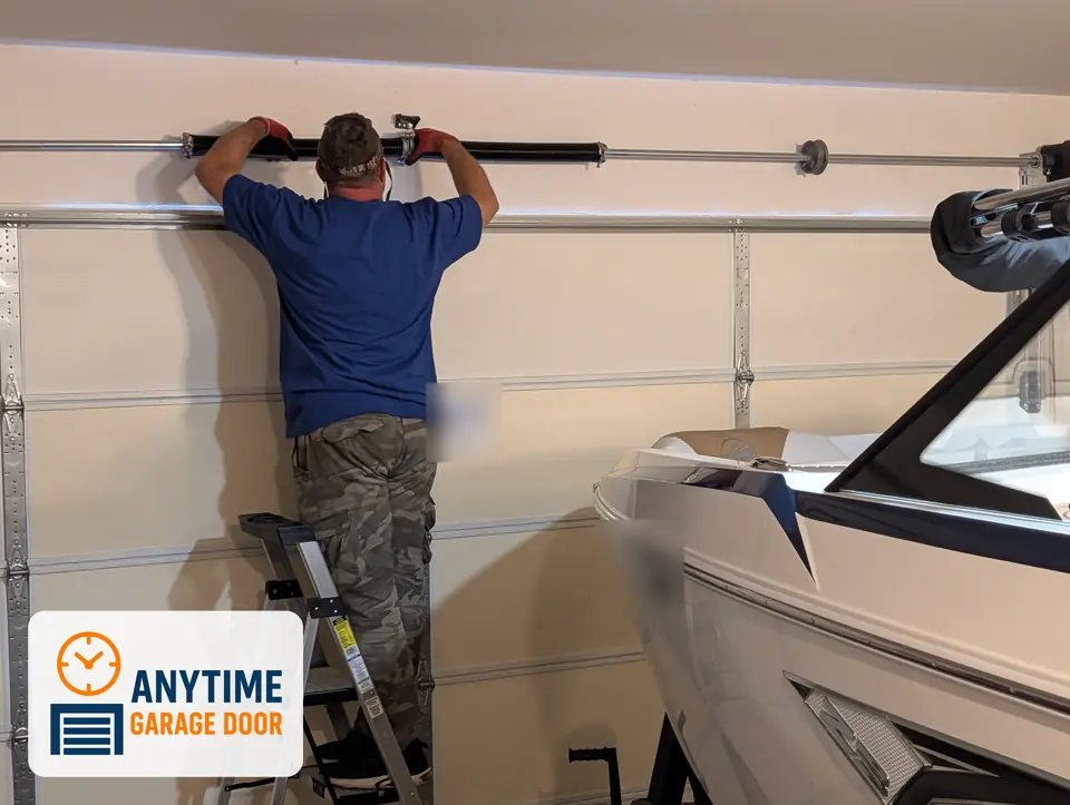 Worker on a stepladder with both hands on a torsion spring above a garage door, with a boat parked beside him